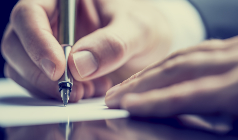 Close up of a hand holding a pen to paper, representing the need to write a will