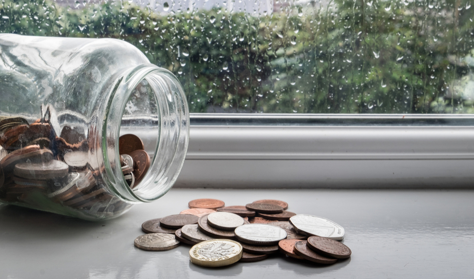 A jar of money tipped over with a rainy day in the background, illustrating an article on emergency savings