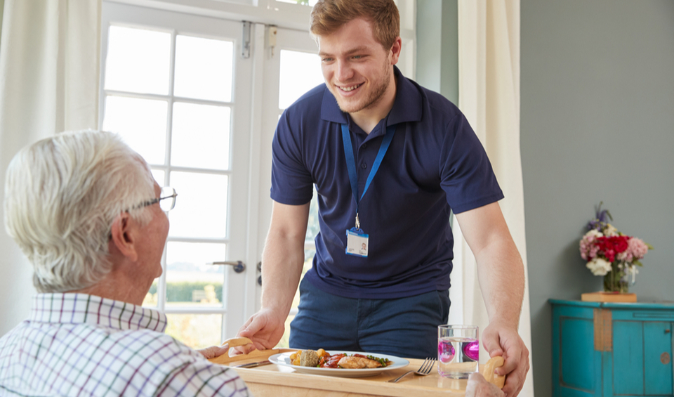 A care worker serving dinner to an older man, illustrating an article on the new care cap
