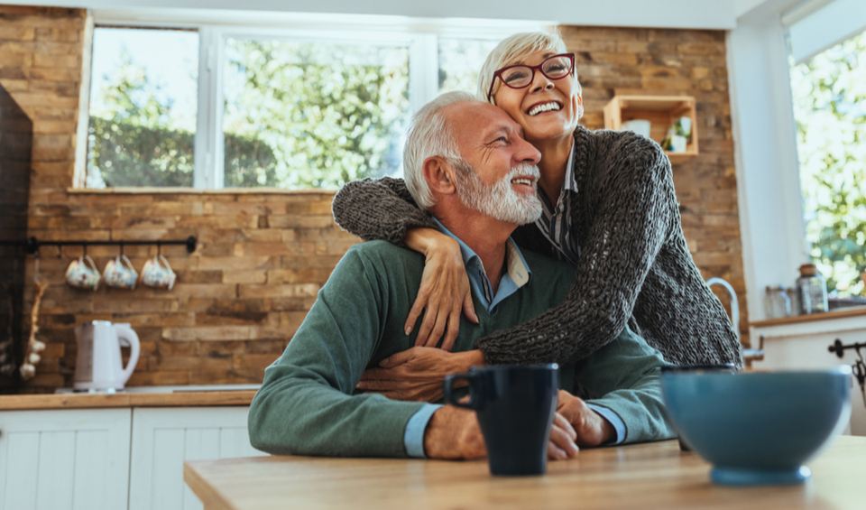 An older couple smiling and hugging at the dinner table, illustrating an article on pension contribution