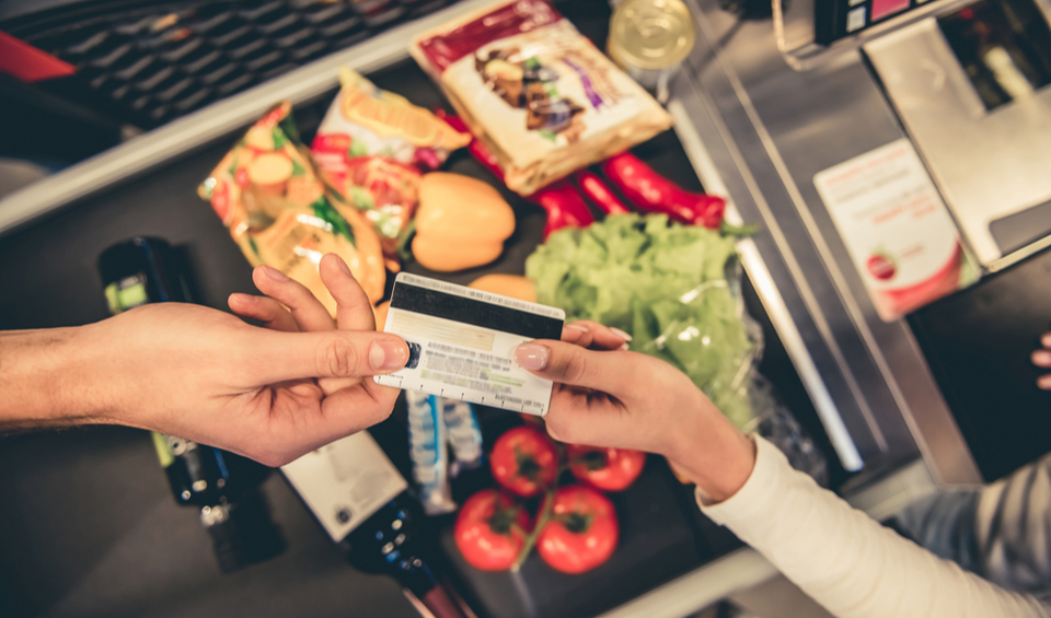 A debit card is held above a supermarket conveyor filled with food, illustrating an article on the cost of living
