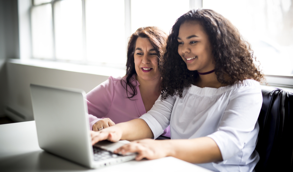 A happy mother and daughter sit at a desk working on a laptop together illustrating an article on the 7 key money lessons to teach children before going to university
